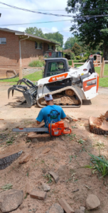 A tree service worker with a chainsaw near a stump and a Bobcat loader at Joel's tree service in Greensboro, NC