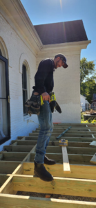 A worker with a tool belt and nail gun standing on the wooden frame of a deck under construction, showcasing building services by Drew's Handy Construction in Charleston, IL.