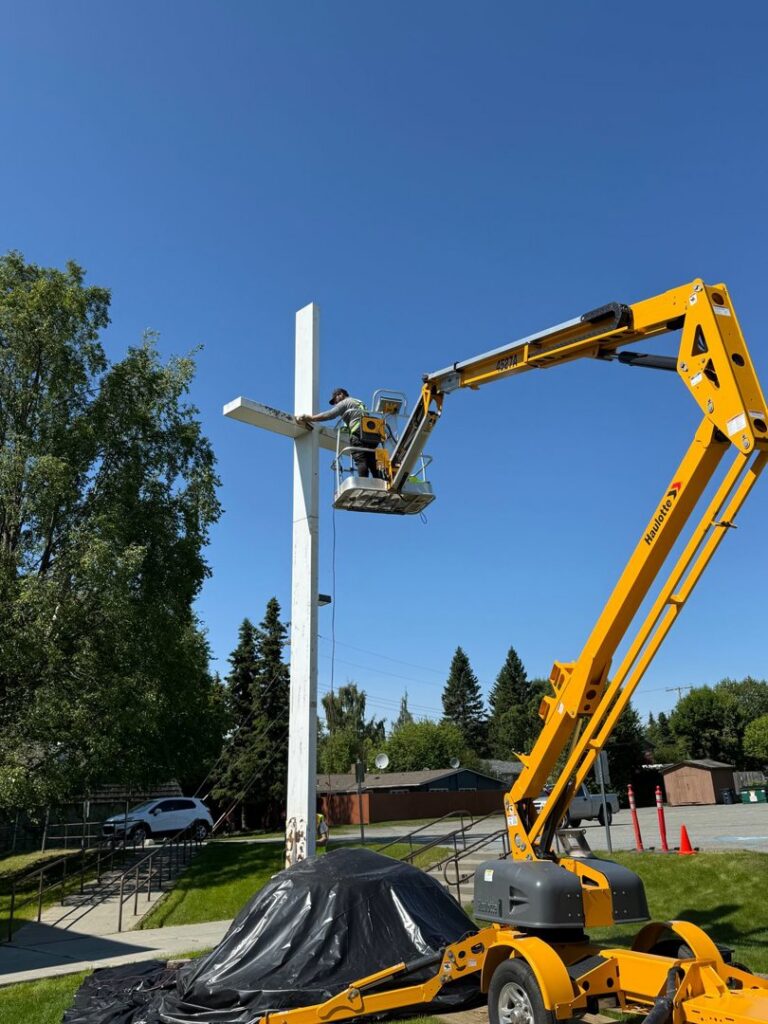 A worker in a lift painting a large white cross structure, a service provided by Alaskan Residential Rescue in Anchorage, AK.