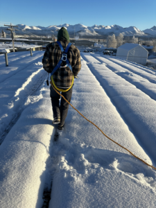 A worker from AK Property Maintenance, Inc. in a safety harness walks on a snow-covered roof in Anchorage, AK.