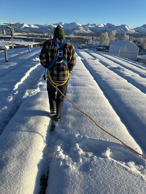 A worker from AK Property Maintenance, Inc. in a safety harness walks on a snow-covered roof in Anchorage, AK.