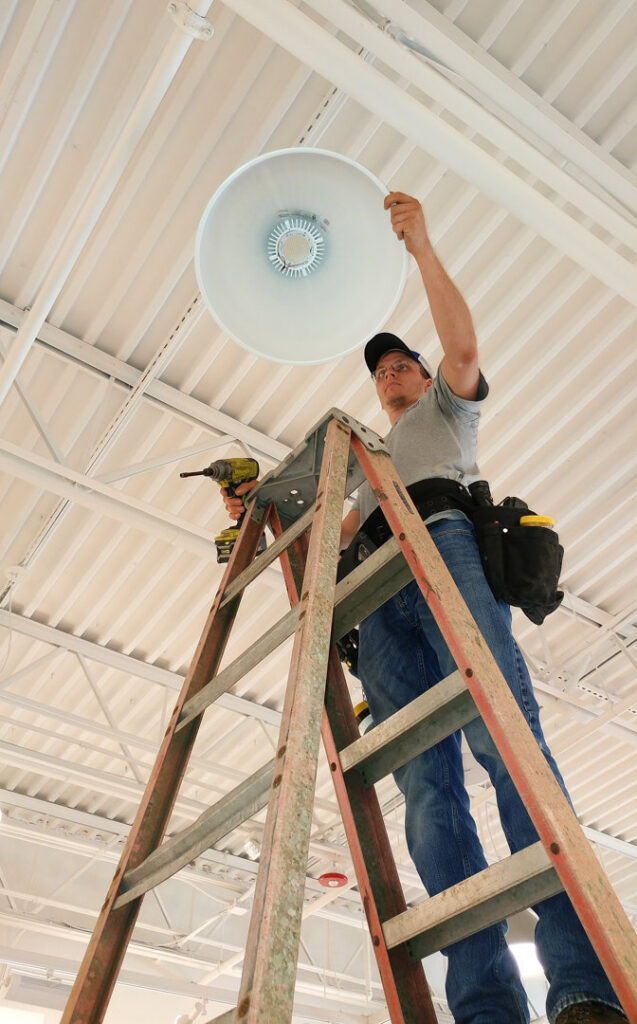 A worker on a ladder installing a large light fixture on a high ceiling for Joyner Electric And Security in Savannah, GA.