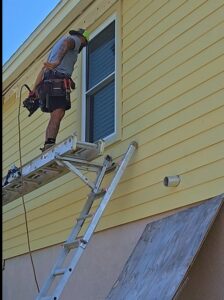 A worker on a ladder installing siding and window trim on a yellow house, performed by Alejandro Melo Contractor corp in Tampa, FL.