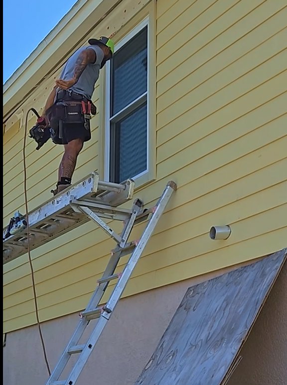 A worker on a ladder installing siding and window trim on a yellow house, performed by Alejandro Melo Contractor corp in Tampa, FL.