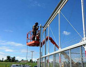 A worker on a lift installing a tall chain link fence with barbed wire for R & C Fence in Fort Wayne, IN.