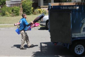 A worker loading various items like bags, chairs, and a blue bucket into a Junk Away & Cleaning trailer in Sacramento, CA.