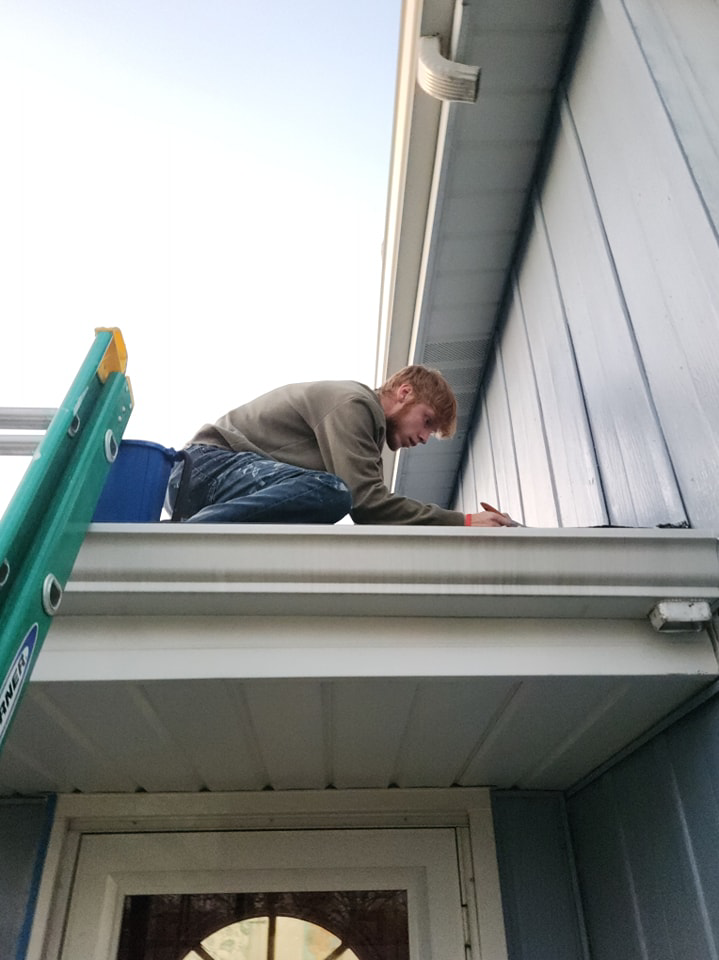 A worker on a ladder carefully painting or sealing the fascia of a house, demonstrating exterior work by Drew's Handy Construction in Charleston, IL.