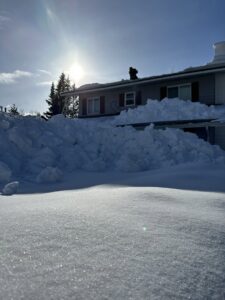 A worker on a roof performing snow removal, with a large pile of cleared snow on the ground, by Alaskan Residential Rescue in Anchorage, AK.