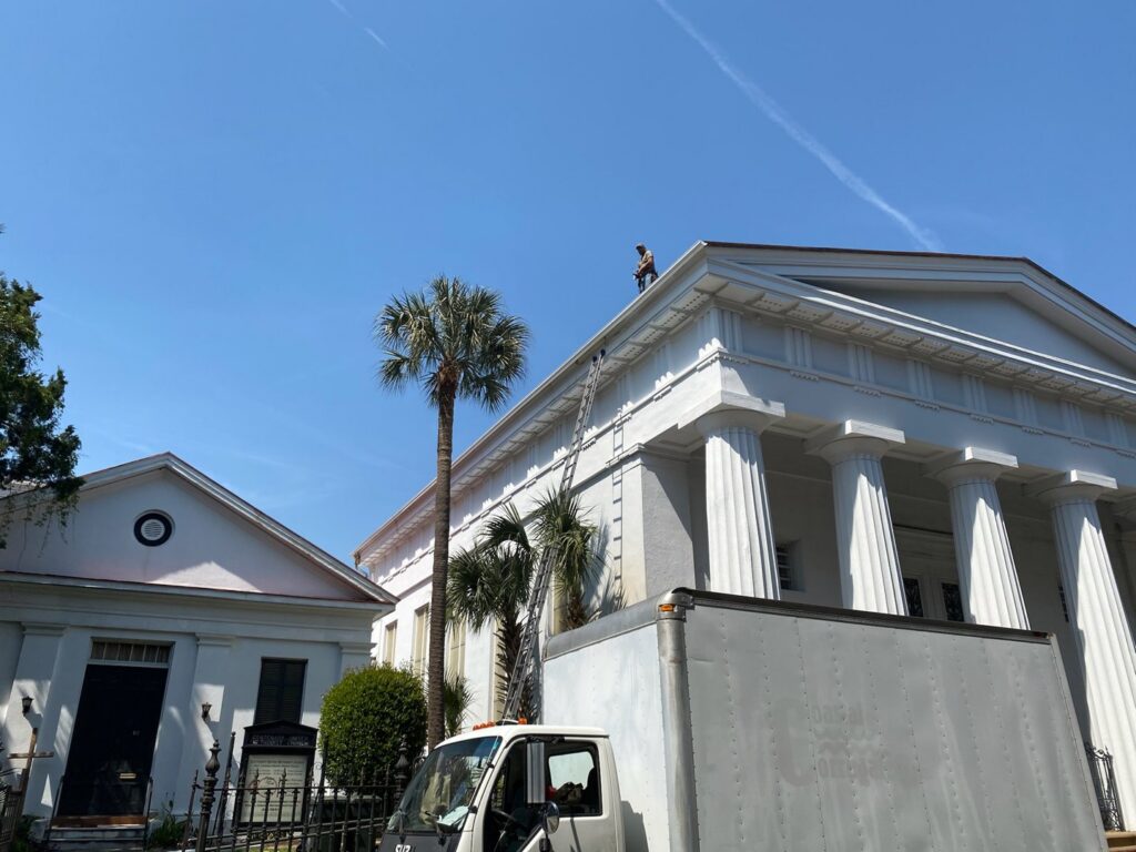 A worker on the roof of a large building with a ladder, performing exterior maintenance for Charleston Exteriors in Charleston, SC.