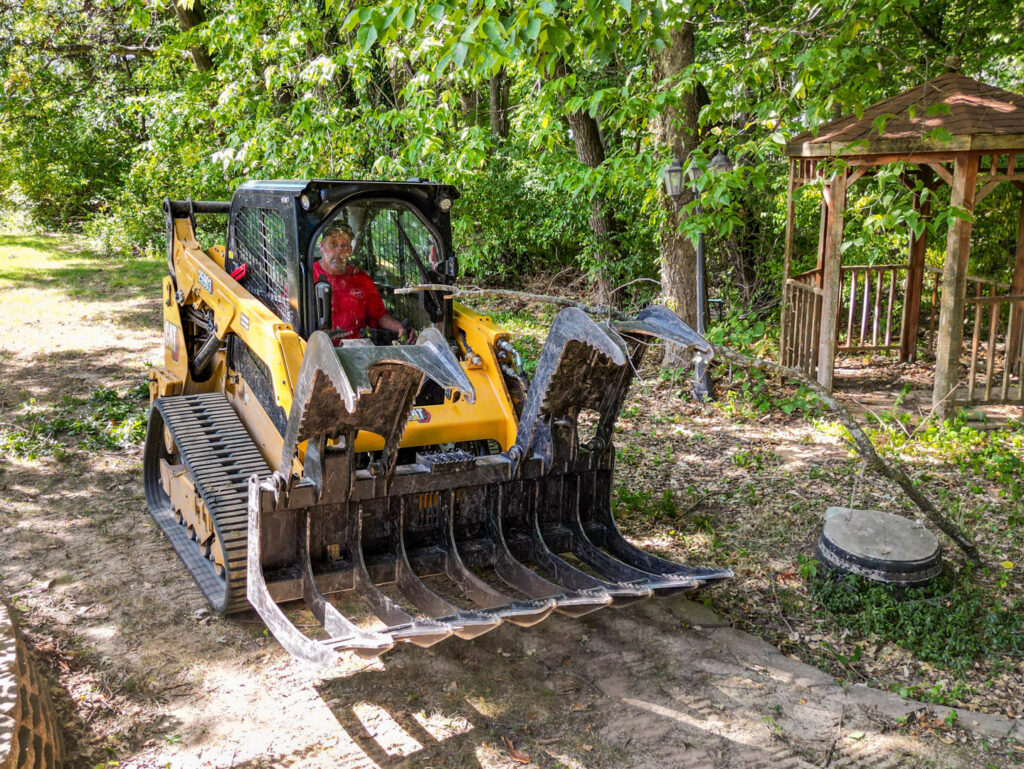 A worker operating a skid steer with a grapple attachment, moving branches for Braik's Tree Care in Columbia, MO.