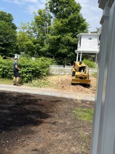 A worker operating a stump grinder next to a white picket fence, performing stump removal for Grin & Grind Stump Removal LLC in Worcester, MA.