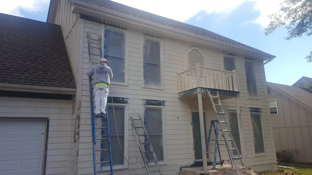 A worker on a ladder painting a house exterior with windows covered for protection by E.L. Construction in Kansas City, MO.