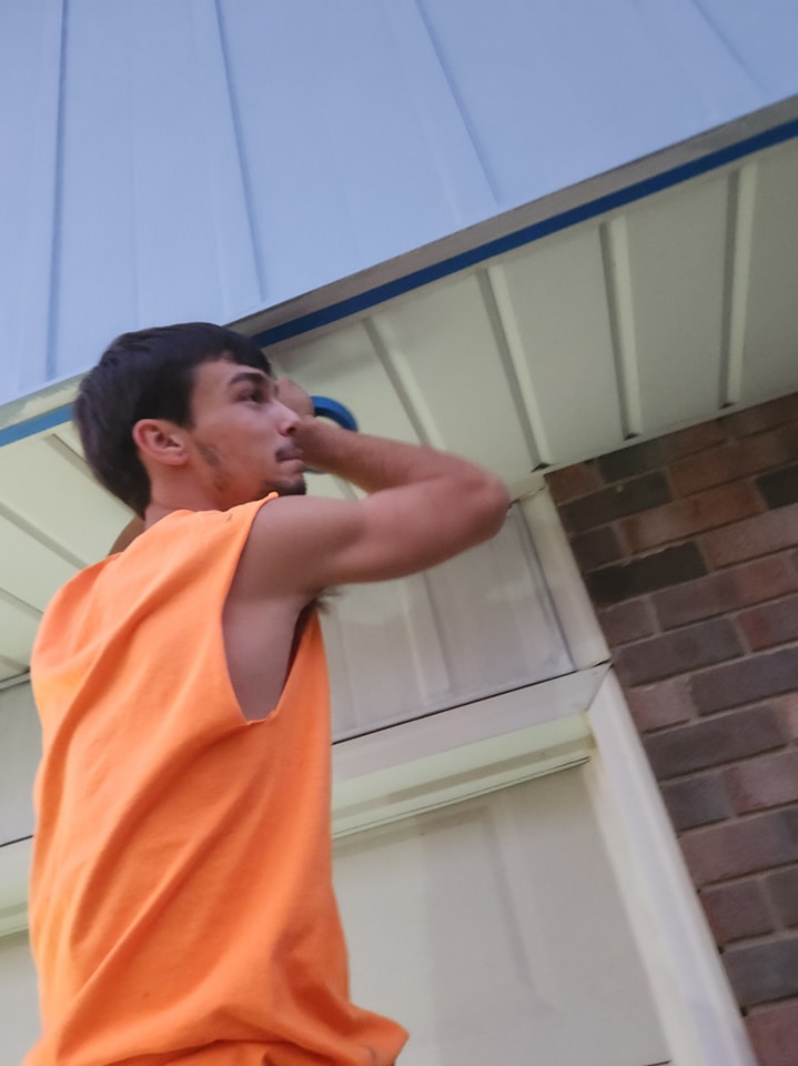 A worker in an orange shirt painting the soffit and eaves of a house, showcasing exterior painting services by Drew's Handy Construction in Charleston, IL.