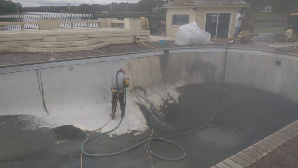 A worker in protective gear pressure washing the inside of an empty swimming pool, a service provided by Vanwin Coatings of Virginia in Chesapeake, VA.