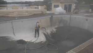 A worker in protective gear pressure washing the inside of an empty swimming pool, a service provided by Vanwin Coatings of Virginia in Chesapeake, VA.