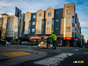 A Bay Area Bin Support worker in a high-visibility vest pushing a large dumpster across a street in San Leandro, CA.