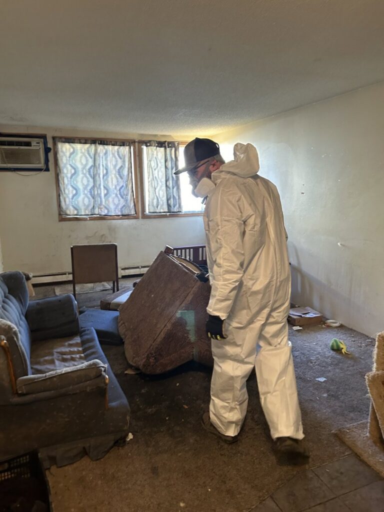 A worker in a protective suit removing a couch from a messy room during a cleanout by The Goat Junk Removal in Ankeny, IA