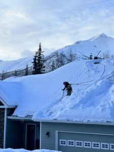 A worker actively removing snow from a residential roof with mountains in the background for Alaskan Residential Rescue in Anchorage, AK.