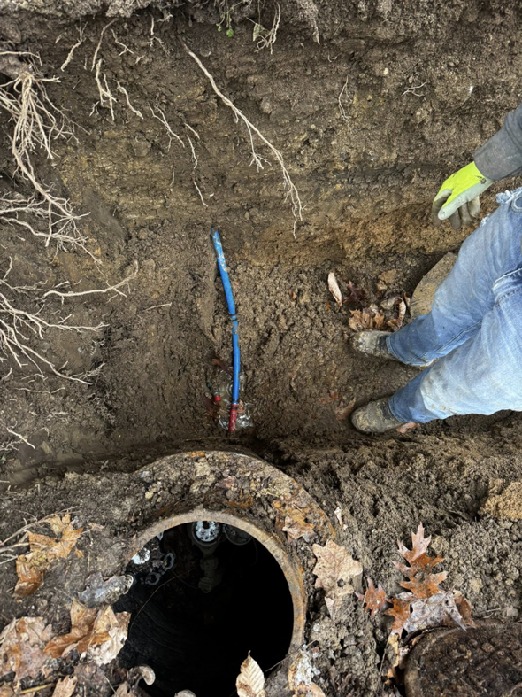 A worker in a trench repairing an underground blue pipe near a manhole, performed by Richardson Ridge Construction in Bloomington, IN.