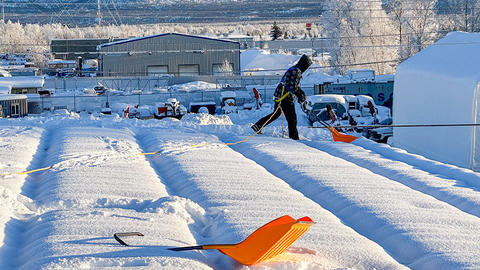 A worker from AK Property Maintenance, Inc. is actively shoveling snow from a roof while wearing safety gear in Anchorage, AK.