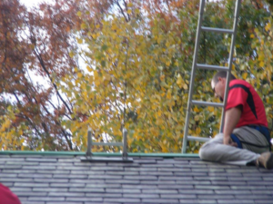 A worker sitting on a slate roof next to a ladder, performing services for Superior Roof Support in Milano, TX.