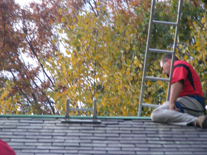 A worker sitting on a slate roof next to a ladder, performing services for Superior Roof Support in Milano, TX.