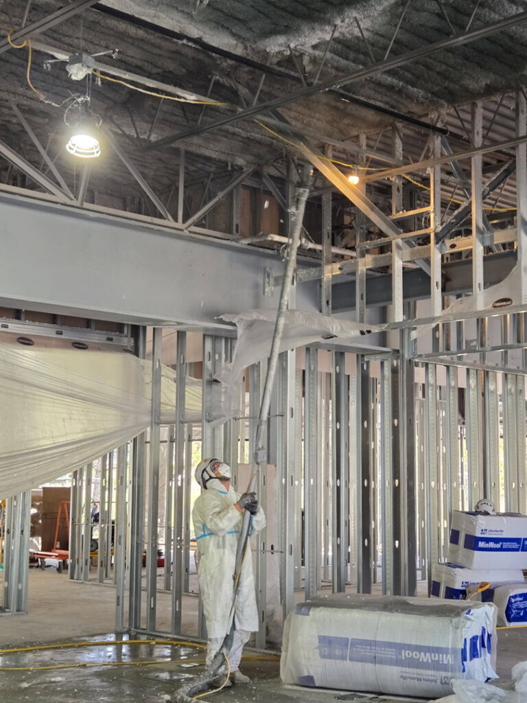 A worker in a protective suit spraying insulation or fireproofing onto a ceiling structure for Bell SPI - Shamrock Painting in Denver, CO.