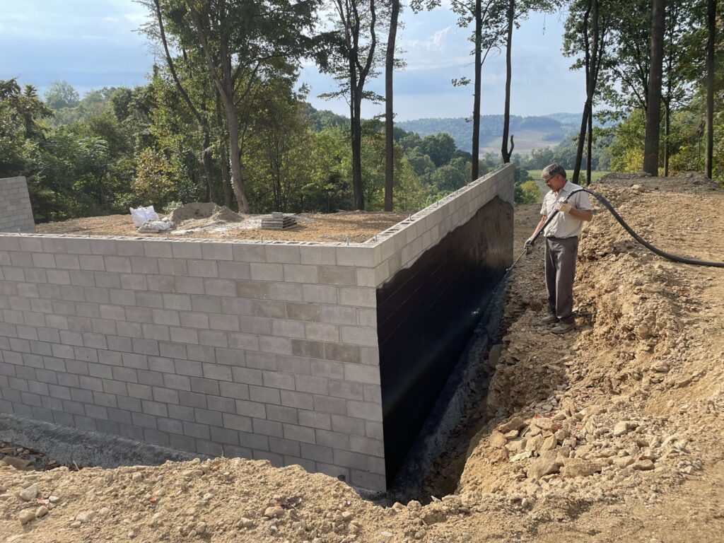A worker spraying a black waterproofing membrane onto a new concrete block foundation for TCM Waterproofing, LLC in Canton, OH.