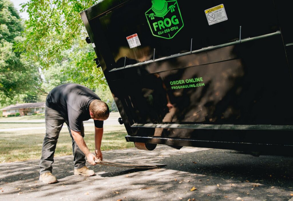 A worker sweeping debris from under a black Frog Hauling dumpster after a junk removal job in Columbus, OH.