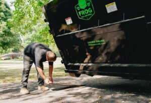 A worker sweeping debris from under a black Frog Hauling dumpster after a junk removal job in Columbus, OH.