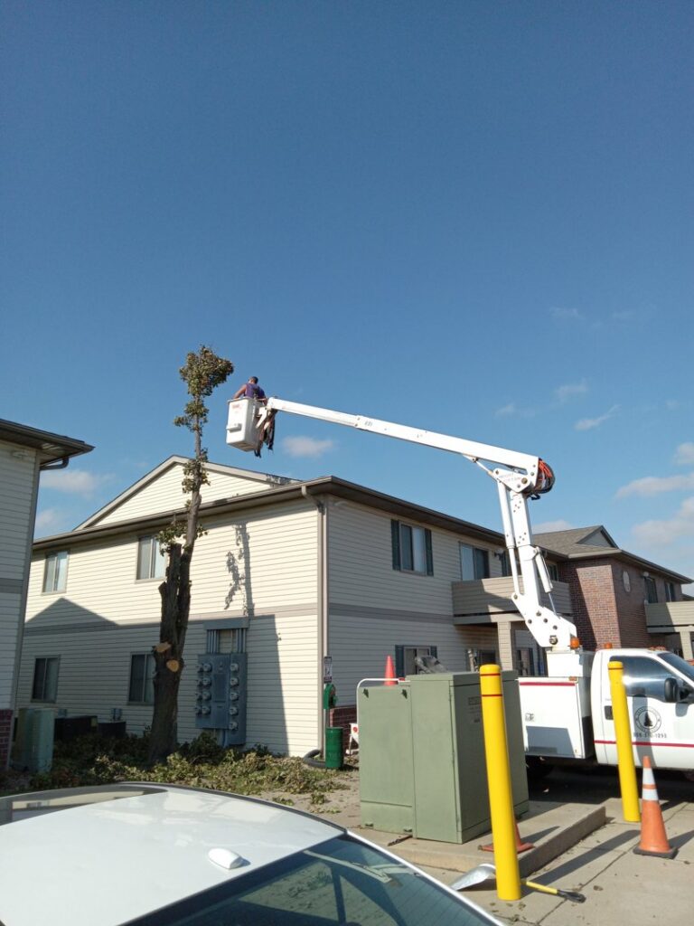 A worker in a bucket truck trimming a tree next to an apartment building for Grand Island Tree Service in Grand Island, NE.