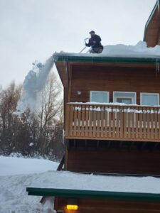 A worker using a snow blower to remove snow from a roof, with icicles visible, for Alaskan Residential Rescue in Anchorage, AK.