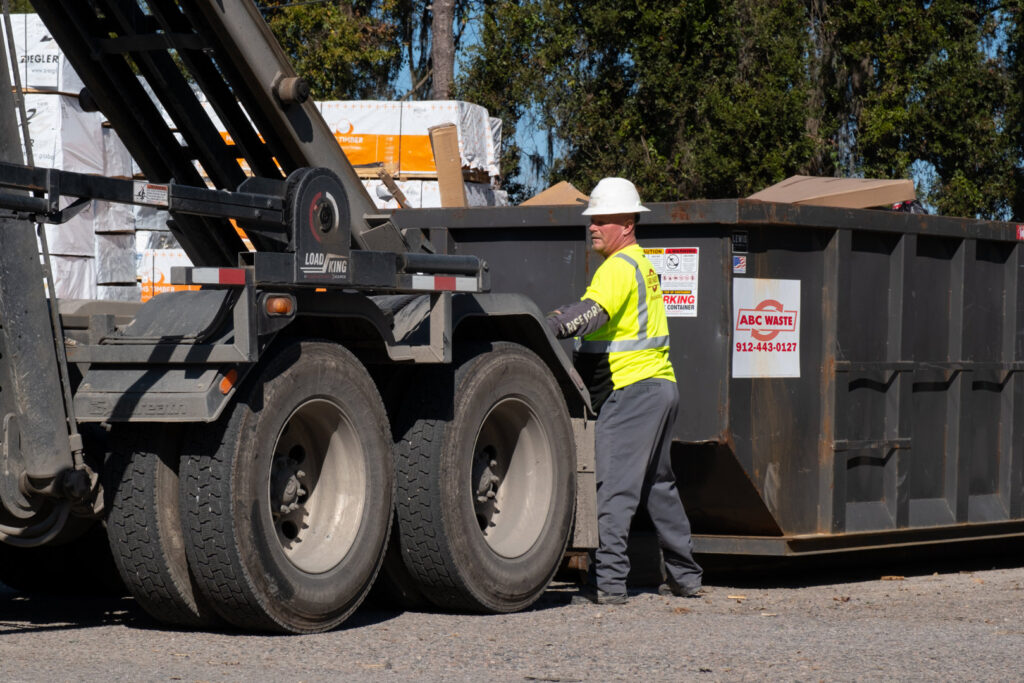 A worker in a hard hat and safety vest standing by a roll-off dumpster from ABC Waste of Savannah, Inc. in Savannah, GA.