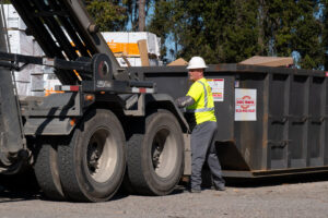 A worker in a hard hat and safety vest standing by a roll-off dumpster from ABC Waste of Savannah, Inc. in Savannah, GA.