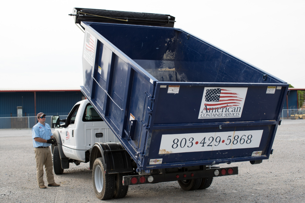 A worker stands next to a truck raising an empty roll-off dumpster for American Container Services, Inc in Columbia, SC.