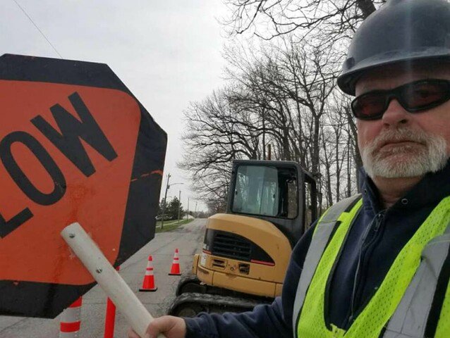 A worker holding a 'SLOW' sign at a job site with an excavator for Hessville Plumbing in Hammond, IN.