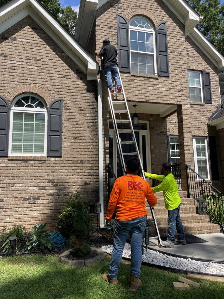 Workers on a ladder performing gutter or fascia repair on a brick house, a service from Roofing Experts Care in Charlotte, NC.