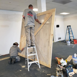 Two workers from Tucker Paint & Construction installing a large wooden barn door, with tools and paint supplies visible, in Fulton, Canton, OH.