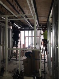 Two workers on ladders installing pipes and conduits in a framed room for Design Build Associates Inc in Baton Rouge, LA.