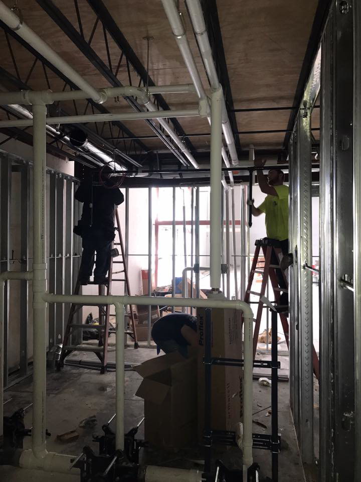 Two workers on ladders installing pipes and conduits in a framed room for Design Build Associates Inc in Baton Rouge, LA.