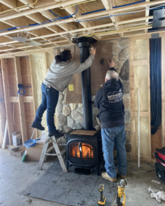 Two workers installing a wood-burning stove and chimney pipe, a service from Spinosa Construction And Maintenance in Tewksbury, MA.