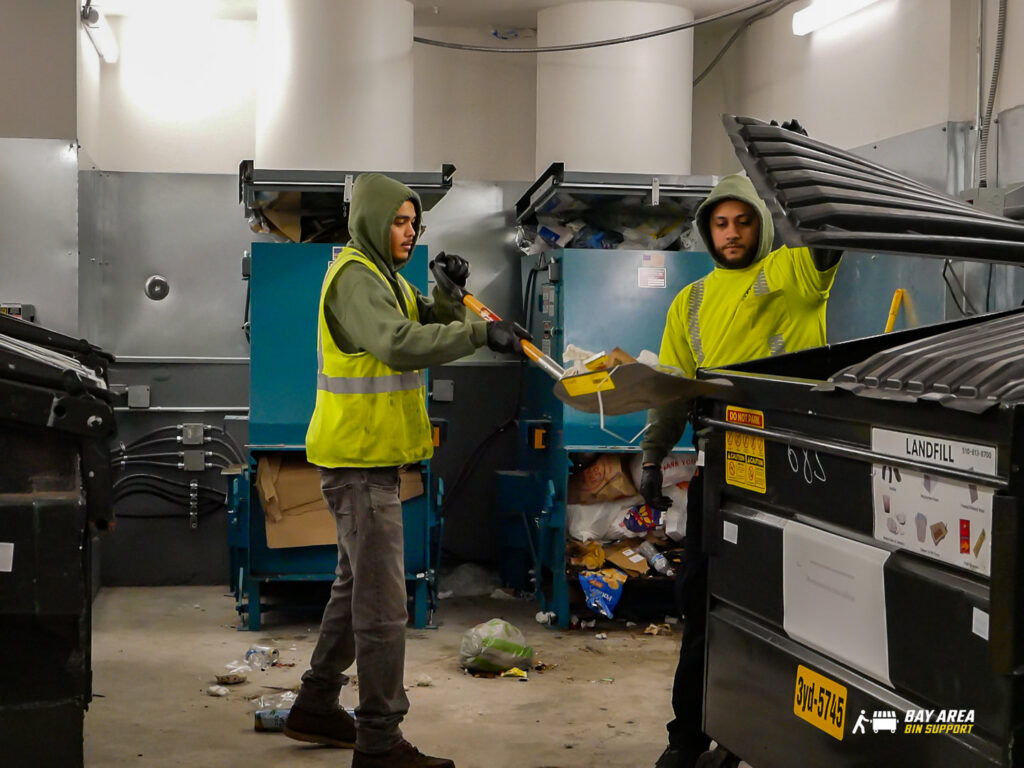 Two Bay Area Bin Support workers loading trash into a dumpster with a shovel in San Leandro, CA.