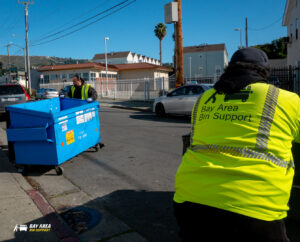 Two Bay Area Bin Support workers moving a blue commercial dumpster on a street in San Leandro, CA.