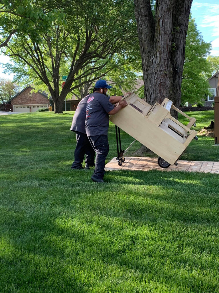 Two Haul-Away Junk Removal workers moving a large wooden cabinet on a dolly across a lawn in Overland Park, KS.
