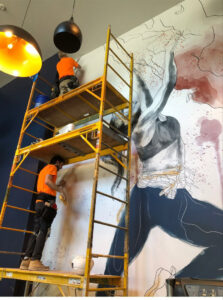 Two workers on scaffolding painting a wall next to a mural for Bell SPI - Shamrock Painting in Denver, CO.