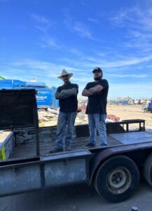 Two Goblins Junk Removal workers standing on a trailer at a junk disposal site, with piles of debris in the background, in Wheat Ridge, CO.