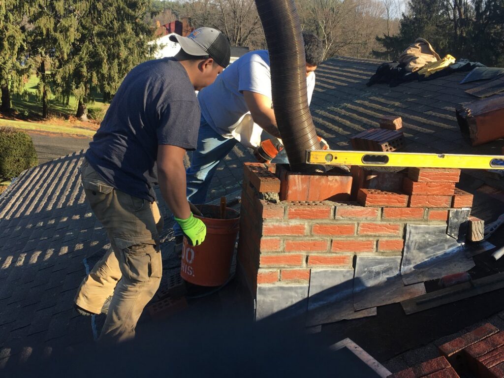 Two workers repairing or rebuilding a brick chimney on a residential roof, a service offered by Advanced Seamless Gutters, Inc. in Chicopee, MA.