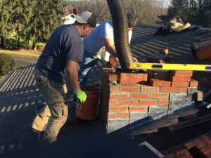 Two workers repairing or rebuilding a brick chimney on a residential roof, a service offered by Advanced Seamless Gutters, Inc. in Chicopee, MA.