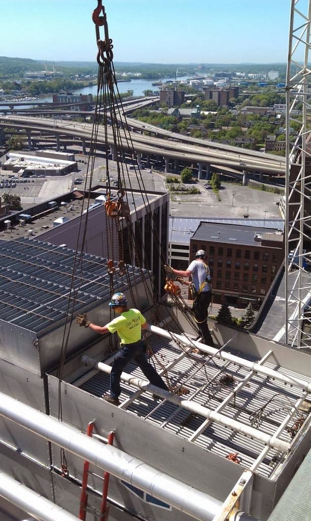 Workers from Mullins Rigging rigging a large rooftop HVAC unit with a city skyline in the background in Green Island, NY