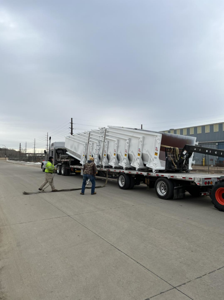 Workers securing new white roll-off dumpsters onto a trailer for Kuchel Roll-Offs in Sioux City, IA.